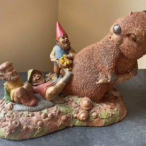 May include: A whimsical figurine of a brown bear with three gnomes. The bear is standing on its hind legs and has its front paws raised. The gnomes are sitting on the ground in front of the bear. The figurine is made of a brown, tan, and green material and is set on a base of brown, tan, and green material.