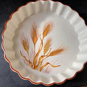 May include: A white ceramic baking dish with a scalloped edge and a brown rim. The dish features a design of wheat stalks in shades of yellow and brown.