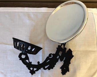May include: A black cast iron wall-mounted soap dish with an oval, silver-colored dish. The soap dish has a decorative, ornate design. The dish is mounted on a white linen surface.