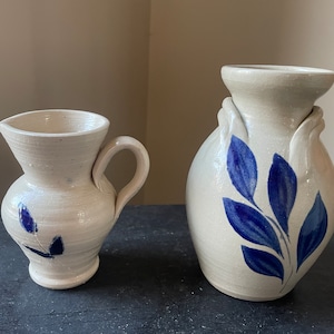 May include: Two ceramic pottery vases with blue and white floral designs. The smaller vase has a handle and is a pitcher. The larger vase has a wider opening and a more rounded shape.