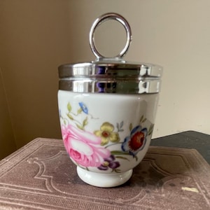 May include: A white porcelain egg cup with a silver lid and a floral design featuring pink, blue, and yellow flowers. The egg cup has a silver handle.