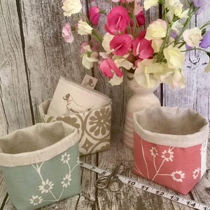 May include: Three fabric storage baskets in light blue, beige, and pink with white floral patterns. The beige basket features a dog illustration. A metal measuring tape shows dimensions in cm, and a pair of metal scissors rests nearby.