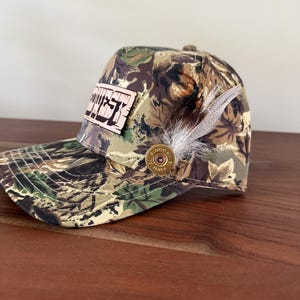 May include: Camouflage baseball cap with a wooden patch that reads "DUST". The cap features a feather and a decorative bullet casing. The hat is designed with a curved brim and a button on top.