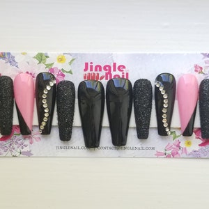 May include: A set of coffin-shaped false nails. The nails feature a variety of designs, including solid black, sparkling black glitter, and pink with a black diagonal accent. Some nails are adorned with a line of rhinestones. The brand name is "Jingle Nail."