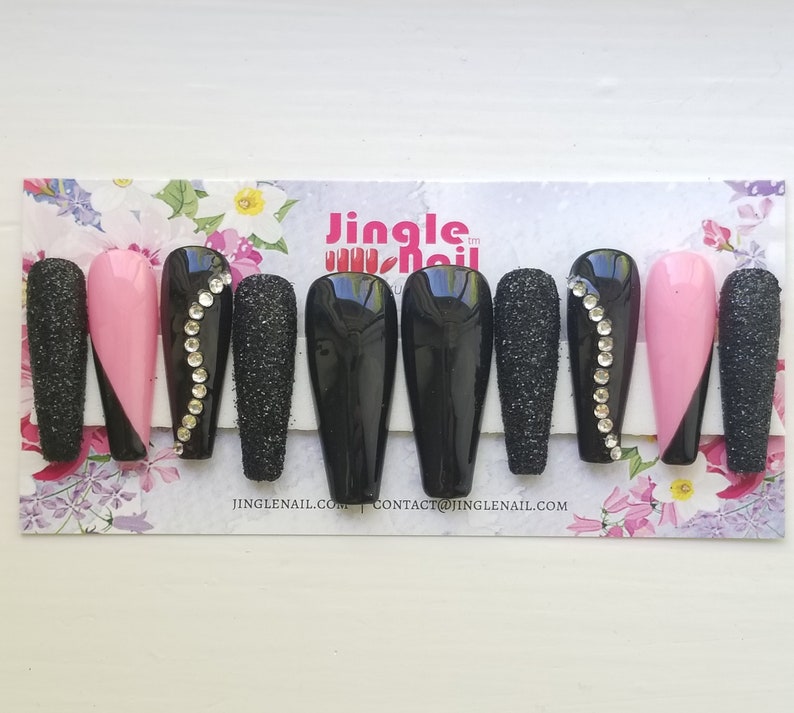 May include: A set of coffin-shaped false nails. The nails feature a variety of designs, including solid black, sparkling black glitter, and pink with a black diagonal accent. Some nails are adorned with a line of rhinestones. The brand name is "Jingle Nail."