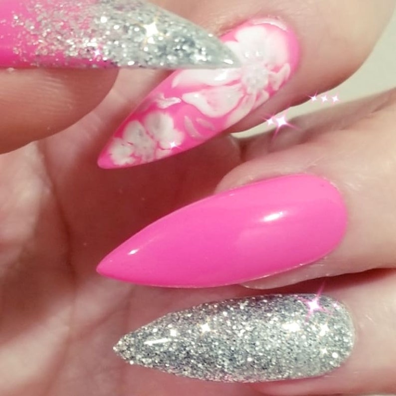 May include: Close-up of manicured nails in a variety of designs. One nail is a solid bright pink, another is covered in silver glitter, and a third features a pink base with white floral accents. The nails are long and pointed.