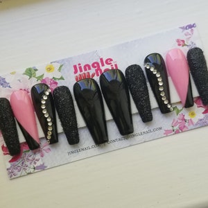 May include: A set of long, coffin-shaped press-on nails. The nails feature a mix of designs, including solid black, glittery black, and pink with a black diagonal accent. Some nails are adorned with a line of rhinestones.