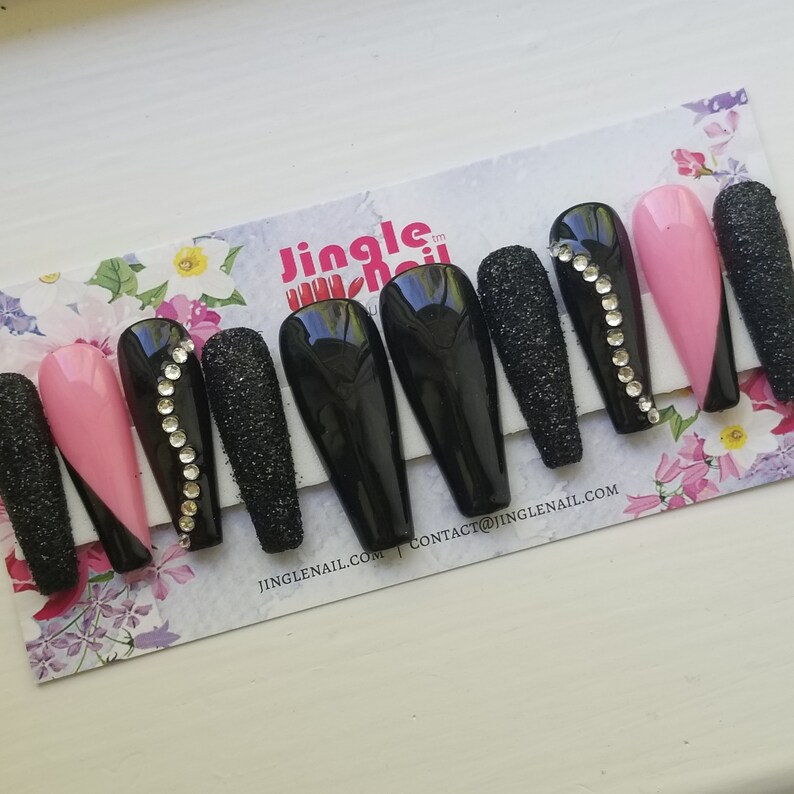 May include: A set of long, coffin-shaped press-on nails. The nails feature a mix of designs, including solid black, glittery black, and pink with a black diagonal accent. Some nails are adorned with a line of rhinestones.