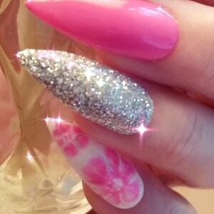 May include: Close-up of three long, pointed artificial nails. One nail is a bright pink, another is covered in silver glitter, and the third is white with a pink floral design. The nails are well-manicured and reflect light.