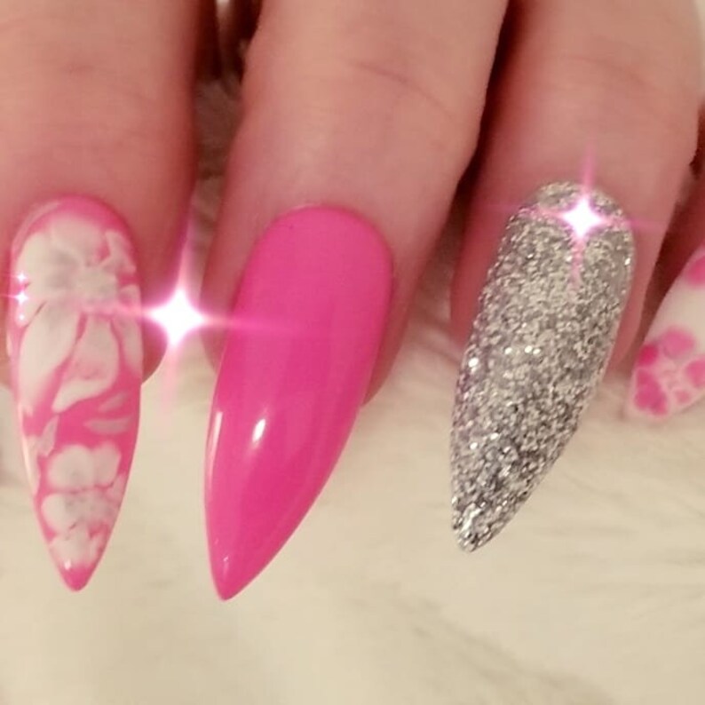 May include: Close-up of a hand with long, pointed acrylic nails. The nails feature a variety of designs, including a bright pink solid color, a silver glitter, and a pink and white floral pattern. The nails are well-manicured and glossy.