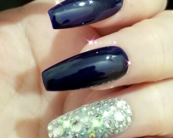 Op de afbeelding: Een hand met lange, coffin-vormige nagels. Vier nagels zijn diep marineblauw gelakt, terwijl één bedekt is met sprankelende zilveren strass-steentjes. De nagels hebben een glanzende afwerking.