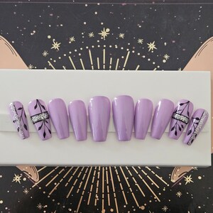 Op de afbeelding: Een set lavendelkleurige valse nagels. Sommige nagels hebben een zilveren glitterontwerp met zwarte bladaccenten. De nagels zijn gerangschikt op een wit oppervlak, met een hemels thema op de achtergrond.
