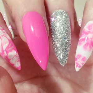 May include: A hand displaying long, pointed artificial nails. The nails feature a vibrant color palette: hot pink, silver glitter, and white with pink floral designs. The nails are a stiletto shape, adding a bold and stylish touch.