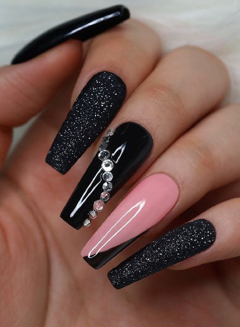 May include: A hand displays long, coffin-shaped artificial nails. The nails feature a variety of designs, including solid black, glitter-covered black, a pink nail, and a black nail with a diagonal pink tip. One nail is adorned with rhinestones.
