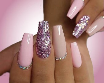 Op de afbeelding: Een hand met lange, coffin-vormige acrylnagels. De nagels zijn geverfd in verschillende tinten roze en nude, met één nagel bedekt met glinsterende paarse glitter. Sommige nagels zijn versierd met kleine, heldere strass-steentjes.