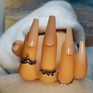 May include: Close-up of a hand wearing several rings and long, nude-colored, coffin-shaped acrylic nails.