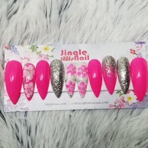 May include: A set of long, pointed artificial nails. The nails feature a vibrant pink color, silver glitter, and floral designs. The nails are arranged on a floral-patterned card with the text "Jingle Nail".