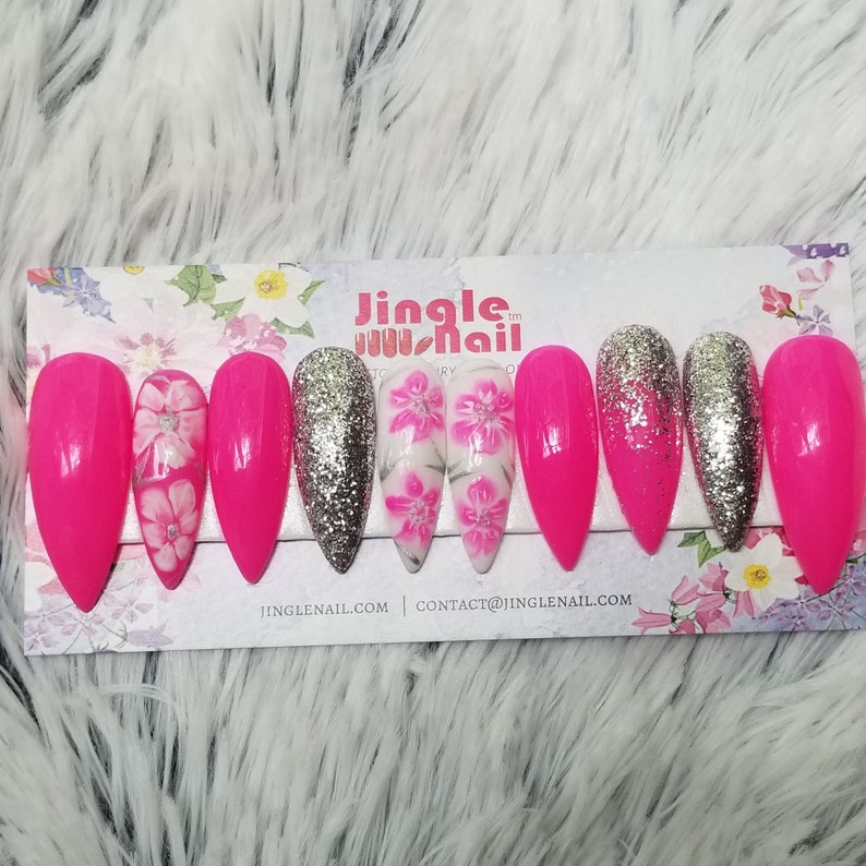 May include: A set of long, pointed artificial nails. The nails feature a vibrant pink color, silver glitter, and floral designs. The nails are arranged on a floral-patterned card with the text "Jingle Nail".