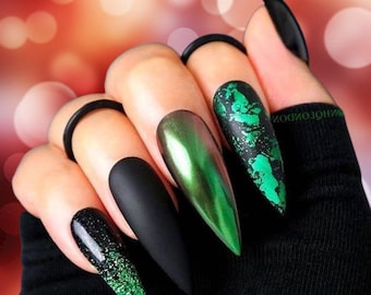 Op de afbeelding: Een hand met lange, puntige kunstnagels. De nagels hebben verschillende ontwerpen, waaronder matzwart, metallic groen, glitter en een groen folie-effect. Een opvallende nagelstyling.