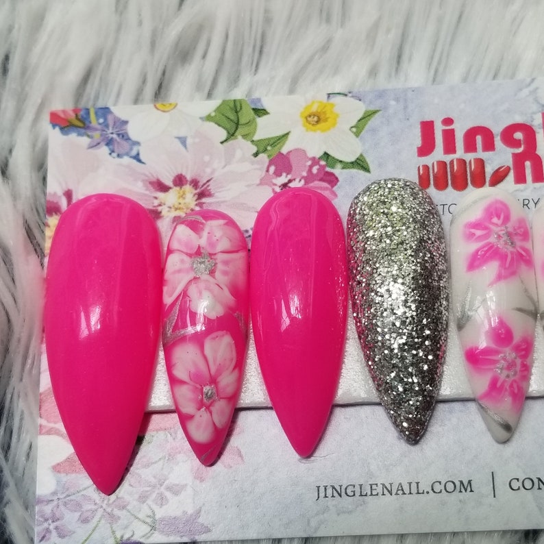 May include: A set of long, pointed artificial nails. The nails are a vibrant hot pink, silver glitter, and white with pink floral designs. The nails are arranged on a floral patterned card.
