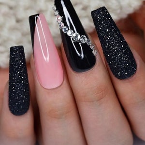May include: Close-up of a hand with long, coffin-shaped acrylic nails. The nails feature a combination of black glitter, glossy black, a light pink, and one nail adorned with rhinestones. The overall design is elegant and stylish.