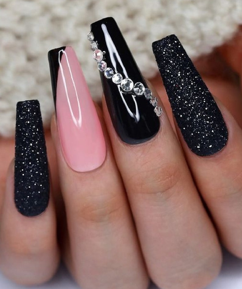 May include: Close-up of a hand with long, coffin-shaped acrylic nails. The nails feature a combination of black glitter, glossy black, a light pink, and one nail adorned with rhinestones. The overall design is elegant and stylish.