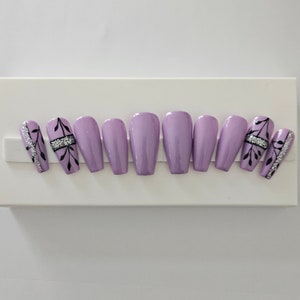 Op de afbeelding: Een set lavendelkleurige persnagels. Twee nagels hebben zilveren glitter- en zwarte bladmotieven. Twee andere nagels hebben een zwart bladmotief met een zilveren strass-accent. De overige nagels zijn effen lavendel.