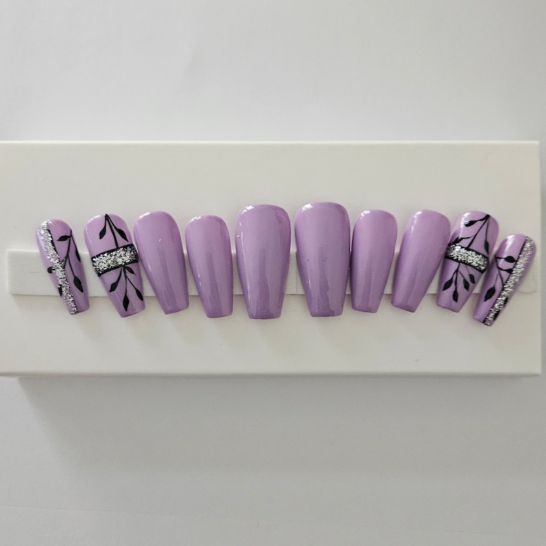 Op de afbeelding: Een set lavendelkleurige persnagels. Twee nagels hebben zilveren glitter- en zwarte bladmotieven. Twee andere nagels hebben een zwart bladmotief met een zilveren strass-accent. De overige nagels zijn effen lavendel.