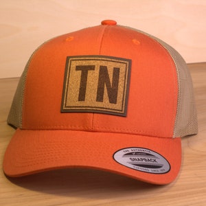 May include: Orange and tan trucker hat with a brown leather patch that says "TN" on it. The hat has a mesh back and a snap closure.