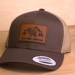 May include: Brown and tan trucker hat with a brown leather patch that says "Great Smoky Mountains" and features a black silhouette of a bear and mountains.