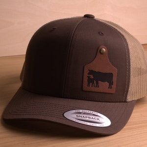 May include: A brown and tan trucker hat with a brown leather patch featuring a silhouette of a cow and calf. The patch has a metal button and the text "The Authentic Snapback" is on the back of the hat.