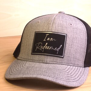 May include: A gray and black trucker hat with a black patch that says "I am Redeemed" in silver lettering.