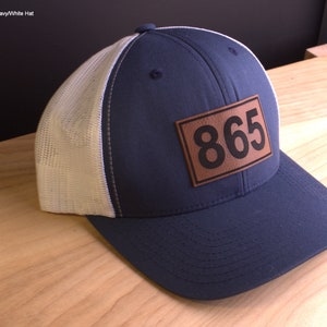 May include: A navy blue and white mesh trucker hat with a brown leather patch that says "865" on it.