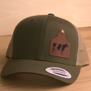 May include: A green and tan trucker hat with a brown leather patch featuring a black and white cow silhouette. The patch is shaped like a cow tag. The hat has the text "The Authentic Snapback" on the back.