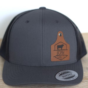 May include: A gray and black trucker hat with a brown leather patch featuring a silhouette of a cow and the text "J.D.W. Cattle Co."