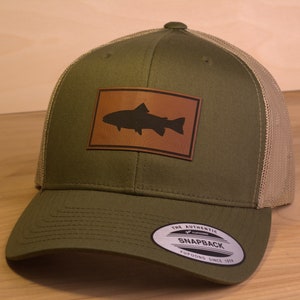May include: A green and tan trucker hat with a brown leather patch featuring a silhouette of a fish. The hat has a mesh back and a curved brim. The hat has a "The Authentic Snapback Yupoong Since 1974" label on the side.