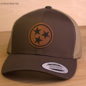May include: Brown and khaki trucker hat with a brown leather patch featuring three stars in a circle. The patch is stitched onto the front of the hat. The hat has a mesh back and a snap closure.