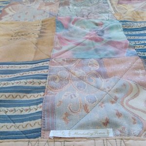 Op de afbeelding: Een patchworkdeken met verschillende kleuren en patronen, waaronder blauw, bruin en beige. De deken is gemaakt van verschillende stoffen en heeft een gestikt ontwerp. De deken heeft een label met de tekst "Made by Tina".