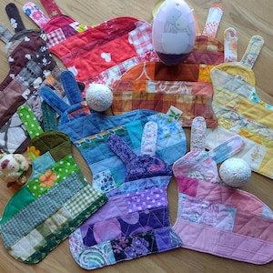 May include: Seven colorful patchwork bunny-shaped coasters with a quilted design. Each coaster features a different color scheme and pattern, including red, blue, green, yellow, and purple. The coasters are made from fabric scraps and are perfect for adding a touch of whimsy to any home.