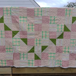 Peut inclure: Une courtepointe en patchwork de couleurs rose, verte et blanche. La courtepointe est composée de carrés et de triangles, et elle a un design simple et élégant.