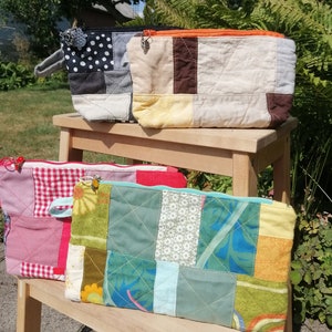 Può includere: Quattro pochette con cerniera in tessuto patchwork di vari colori e motivi, tra cui gingham rosso e bianco, fiori verdi e blu, pois neri e bianchi e marrone e beige. Le pochette sono esposte su uno scaffale di legno.