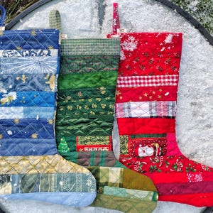 Può includere: Tre calze di Natale fatte di tessuto patchwork. Le calze sono blu, verdi e rosse con vari motivi. Le calze sono appese su una superficie bianca con la neve.