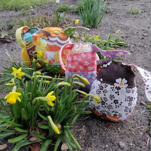 Peut inclure: Quatre œufs de Pâques en tissu colorés avec différents motifs, notamment des motifs floraux, géométriques et rayés. Les œufs sont posés sur un lit de terre brune avec des jonquilles jaunes qui poussent au premier plan.