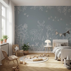 May include: A child's bedroom with a blue-grey wall featuring white line drawings of deer, flowers, and bees. The room includes a white bed, wooden chairs, a round rug with a toy train set, and potted plants. The floor is made of wood.