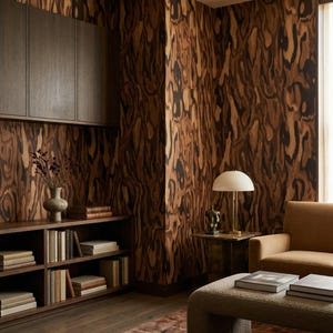 May include: A room interior featuring walls covered in dark brown and black wood grain patterned wallpaper. The space includes a dark wood bookshelf with books, a beige armchair, a textured ottoman, and a table lamp with a white dome shade.