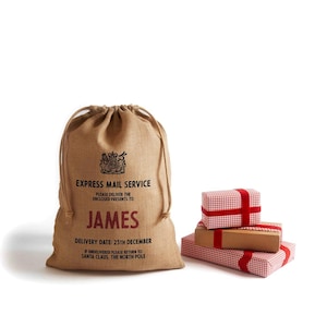 May include: A tan burlap Santa sack with a drawstring closure, featuring the text "EXPRESS MAIL SERVICE" and the name "JAMES" in red. The delivery date is December 25th. Beside the sack are wrapped gifts in red and white checkered paper.