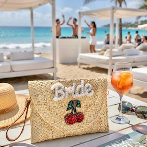 May include: A natural woven straw clutch bag with the word "Bride" in white beads and a cherry design. The bag sits on a white table with a cocktail, sunglasses, and a magazine. The background shows a beach scene.