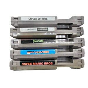 Pode incluir: Uma pilha de cinco cartuchos de jogos cinzentos do Nintendo Entertainment System (NES). Os r&oacute;tulos incluem "CAPTAIN SKYHAWK", "KONAMI", "Shadowgate", "SPY HUNTER" e "SUPER MARIO BROS.". Os cartuchos est&atilde;o empilhados.