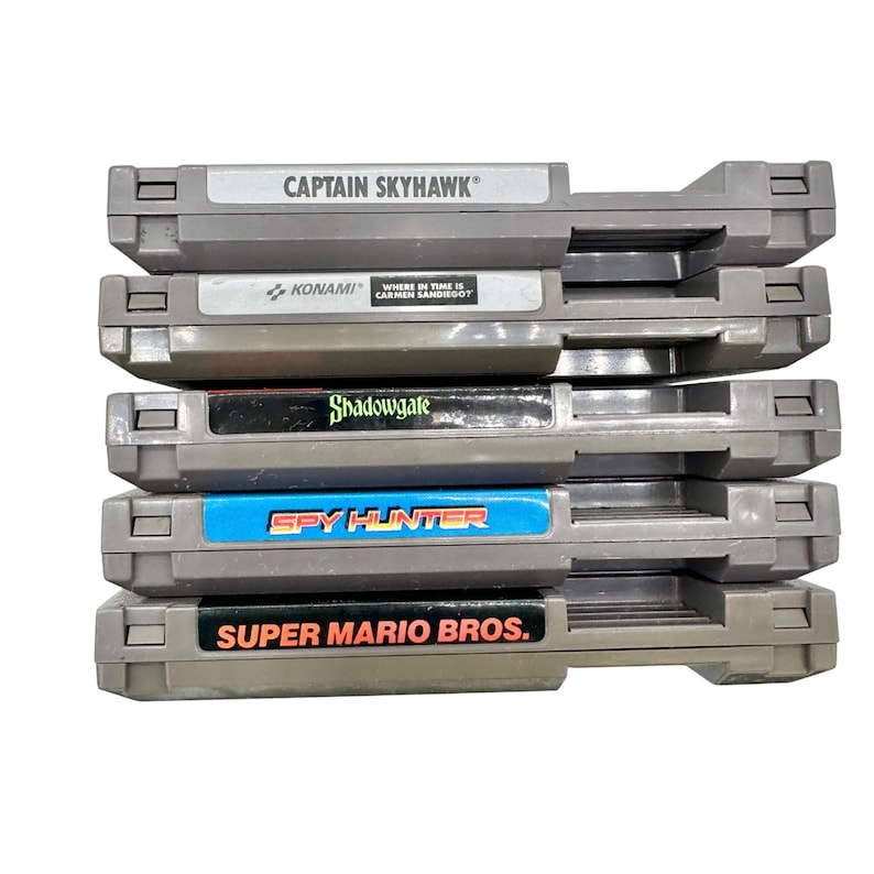 Pode incluir: Uma pilha de cinco cartuchos de jogos cinzentos do Nintendo Entertainment System (NES). Os r&oacute;tulos incluem "CAPTAIN SKYHAWK", "KONAMI", "Shadowgate", "SPY HUNTER" e "SUPER MARIO BROS.". Os cartuchos est&atilde;o empilhados.
