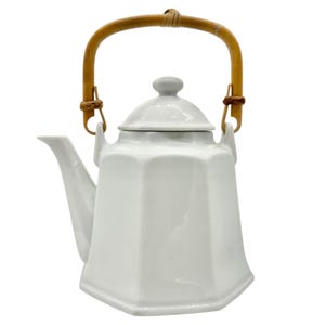 Könnte beinhalten: Eine weiße Keramik-Teekanne mit geometrischem Design und Bambusgriff. Die Teekanne hat einen Deckel mit kleinem Knopf und einen Ausguss zum Ausgießen. Der Griff ist mit geflochtenem Bindfaden befestigt. Die Teekanne ist ein klassisches Design.